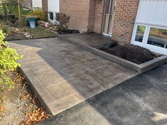 Stamped concrete walkway and porch with rectangular pattern beside a raised garden bed filled with soil at a brick house entrance.
