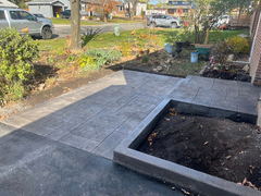 Stamped concrete walkway and patio next to a raised garden bed with fresh soil, surrounded by landscaping and a residential street.