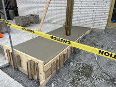 Freshly poured concrete walkway with wooden forms and caution tape in front of a brick building.
