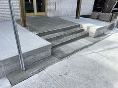 Freshly poured concrete steps and ramp leading to a building entrance with a brick wall and gold double doors.