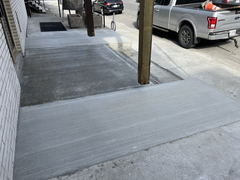 Freshly poured concrete sidewalk with brushed finish adjacent to a building and a pickup truck parked on the street.