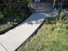 Newly poured concrete walkway with subtle brushed finish leading to porch steps, bordered by garden beds and flowers.