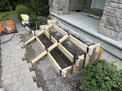Wood forms with steel rebar set up for concrete porch steps construction next to stone pavers and landscaping.