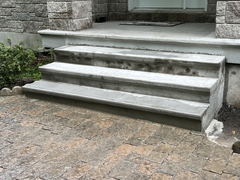 Freshly poured concrete front steps leading to a porch, bordered by stone walls and connecting to a textured paver walkway.