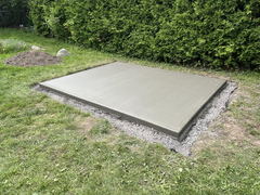 Freshly poured concrete slab foundation with smooth finish, surrounded by gravel and grass in a backyard.