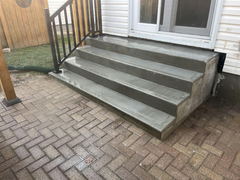 Freshly poured concrete steps with metal railing next to a brick paver patio outside a house.