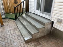 Freshly poured concrete steps with a black metal railing leading to a patio door, bordered by interlocking brick pavers.