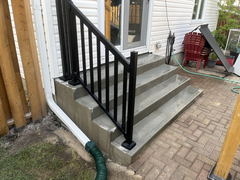 Freshly poured concrete steps with black metal railing leading to a patio door, adjacent to a brick paver walkway and gutter downspout extension.