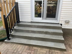 Freshly poured concrete steps with a black metal railing leading to a patio door, adjacent to a brick paver walkway.