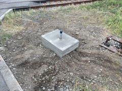 Freshly poured concrete footing with a conduit stub in the center, surrounded by dirt and gravel near railroad tracks.