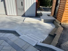 Freshly poured concrete patio with angled steps, retaining walls, and adjoining walkway between metal and log-sided buildings.