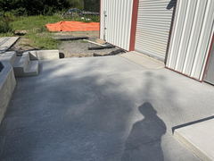 Freshly poured concrete patio with steps leading to a metal building with a roll-up door, gravel area, and orange tarp in the background.