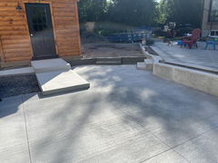 Freshly poured brushed concrete patio and steps leading to a log cabin door, adjacent to unfinished landscaping and a paved seating area.