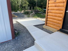 Freshly poured concrete walkway with steps next to wood-sided building and gravel driveway