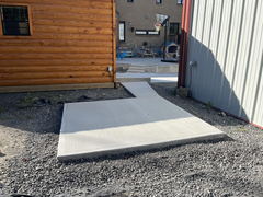 Freshly poured concrete walkway and pad between a wooden building and a metal-sided structure, surrounded by gravel.