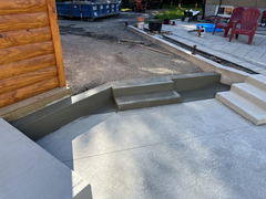Freshly poured concrete walkway and steps with a textured finish next to a log cabin wall and patio area.