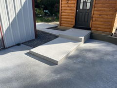 Freshly poured concrete walkway with ramp and step leading to a black door on a log cabin exterior, adjacent to a metal building.