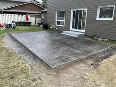 Freshly installed stamped concrete patio with a slate pattern next to a house, bordered by gravel and unfinished landscaping.
