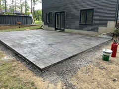 Freshly installed stamped concrete patio beside a modern black house, with surrounding gravel edge and construction materials nearby.