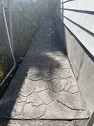 Stamped concrete walkway beside a house with a textured stone pattern and shadow from a fence.