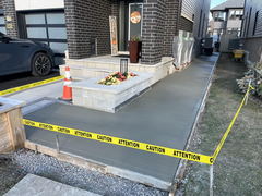 Freshly poured concrete walkway and porch steps with caution tape and traffic cone in front of a modern house entrance.