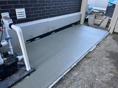 Freshly poured concrete slab next to a building wall with visible pool pump equipment and plumbing on the left.