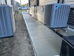Freshly poured brushed concrete walkway alongside a house with several outdoor HVAC units and protective plastic sheeting on the wall.