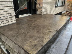 Stamped concrete porch in front of a brick house with wet surface and black door entrance.