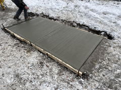 Freshly poured concrete walkway section with wooden forms in a snowy outdoor setting and a worker nearby.