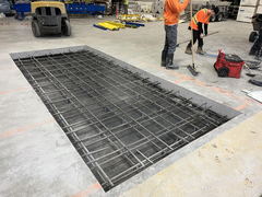 Concrete floor cutout with exposed rebar grid reinforcement, construction workers sweeping nearby in an industrial setting.