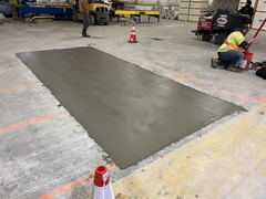Freshly poured concrete patch in a warehouse floor with construction worker nearby and safety cones marking the area