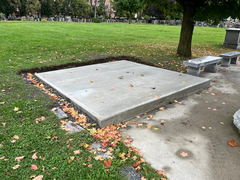 Freshly poured concrete slab surrounded by grass and fallen autumn leaves, next to stone benches in a park setting.