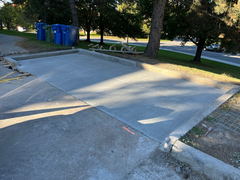 Freshly poured concrete driveway slab with framing boards along the edges, set beside a street with trees and recycling bins in the background.
