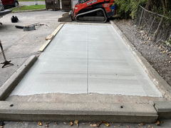 Freshly poured concrete driveway section with control joints, surrounded by old curbs, compact track loader parked at the end