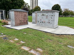 Columbarium structures with engraved plaques and floral tributes on a newly poured concrete walkway in a cemetery.