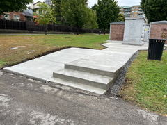 Freshly poured concrete walkway with three steps leading to a columbarium in a cemetery, bordered by grass and a gravel edge.