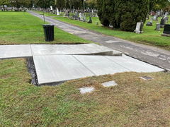 Freshly poured concrete walkway with a step transition, surrounded by grass and gravestones in a cemetery.