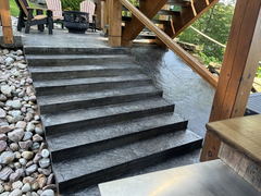 Stamped concrete steps beside a wooden deck with Adirondack chairs and a fire pit, bordered by decorative river rocks.