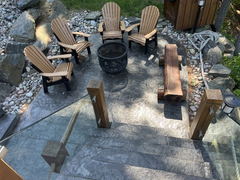 Stamped concrete patio with Adirondack chairs, a fire pit, log bench, glass railing, and surrounding river rocks and boulders.
