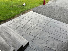Freshly sealed stamped concrete patio with textured pattern next to concrete steps, gravel driveway, and green lawn.