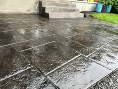Wet stamped concrete patio with a square tile pattern next to concrete steps and green grass.