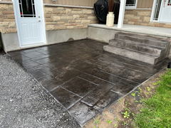 Dark stamped concrete patio with rectangular pattern next to front porch steps and stone veneer house exterior.