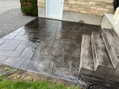 Freshly sealed stamped concrete patio with a textured finish beside concrete steps and a stone wall exterior.