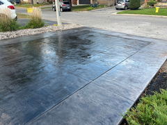 Freshly installed black stamped concrete driveway with a glossy finish adjacent to a landscaped border of rocks and grass.