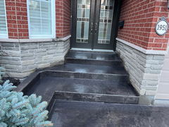 Dark stamped concrete steps and walkway leading to a modern double front door with glass panels on a brick house with stone veneer accents.