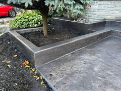 Stamped concrete patio with raised concrete planter box surrounding a tree, adjacent to a stone house exterior.